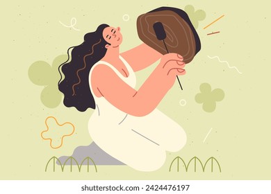Woman shaman plays sacred drum, demonstrating traditional ritual for summoning spirits or attracting good luck. Hippie girl is fond of shamanism and holds shaman drum, sitting on lawn in park.