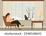 Woman sitting in a chair by the window drinks coffee, a cat is sitting on the windowsill, the weather is bad outside and it’s snowing. Winter weekend at home.
