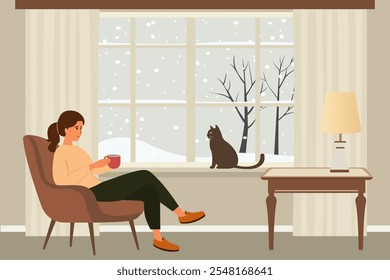 Woman sitting in a chair by the window drinks coffee, a cat is sitting on the windowsill, the weather is bad outside and it’s snowing. Winter weekend at home.