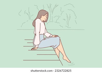 Woman is sitting on pier, lowering into water and enjoying silence and good summer weather. Dreamy young girl looks at river or lake relaxing on pier in public park after hard day work.