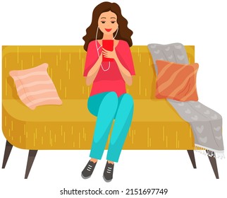 Woman sitting on sofa holds smartphone in her hand. Casual lady in headpones listening to music and browsing social media on mobile device. Girl on couch uses phone for chatting and surfing internet