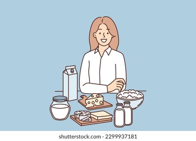 Woman is standing near table with dairy products and yogurt or cheese high in lactose and nutrients. Girl nutritionist offers to eat natural organic dairy products to support health