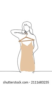 woman stands in front of mirror and leans against hanger with short beige dress with spaghetti straps - one line drawing vector. concept of trying on new dress 