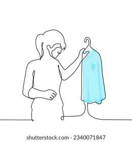 woman stands holding blue shirt on hanger - one line drawing vector. the concept of shopping, trying on in the wardrobe