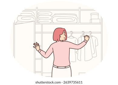Woman stands near large wardrobe closet choosing outfit for going to work and hanging clothes on hanger. Girl is located with back to screen looking at clothes hanging in closet or on counter store