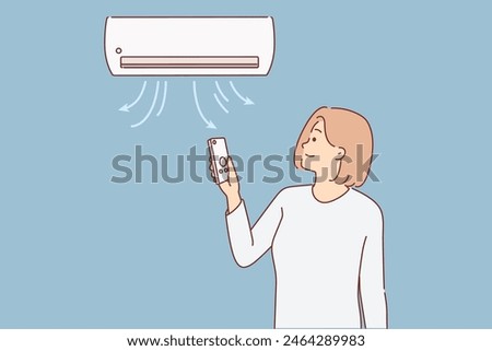 Woman stands under air conditioner and uses remote control to switch operating mode or change temperature. Working conditioner hangs on wall of room, near girl enjoying coolness and fresh air