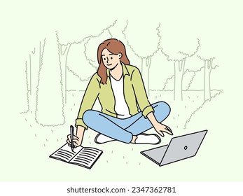 Woman student is sitting on lawn making notes in notebook and using laptop enjoying working in park. Happy girl student watching educational video and writing notes located in park