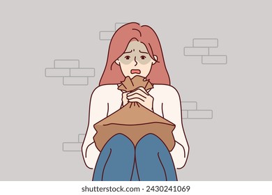 Woman suffering from panic attack holds paper bag for hyperventilation and restoration of normal psychological state. Girl sitting on floor near brick wall and experiencing stress due to panic attack