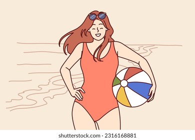 Woman in swimsuit with ball for beach volleyball stands on seashore enjoying summer vacation in tropical resort. Girl tourist smiles posing on beach near ocean and invites you to go on joint tour