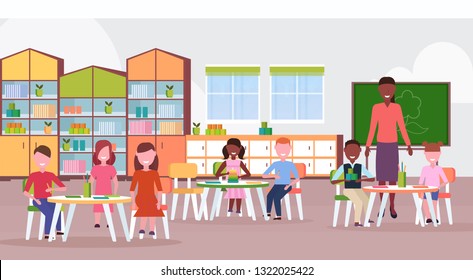 woman teacher teaching mix race boys and girls preschool modern kindergarten children classroom with chalkboard desks chairs kid room interior full length flat horizontal