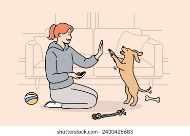 Woman trains dog, sitting on floor near sofa and giving food to puppy after following command. Smart dog stands on two legs, fulfilling requirements of female trainer who enjoys communicating with pet