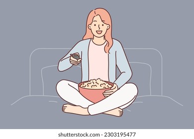 Woman with TV remote turns on series sitting on bed in bedroom and holds cup of popcorn. Girl using TV remote enjoying watching feature film or evening show on cable or sputnik television