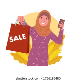 a woman wearing a hijab smiles and shows a paper bag that says sale while holding her cellphone.