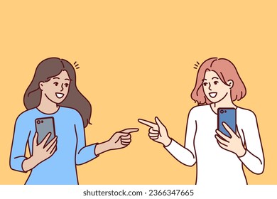 Women with mobile phones point fingers at each other, rejoicing at first meeting after acquaintance in social networks. Happy girls with phones, advertise applications for communication