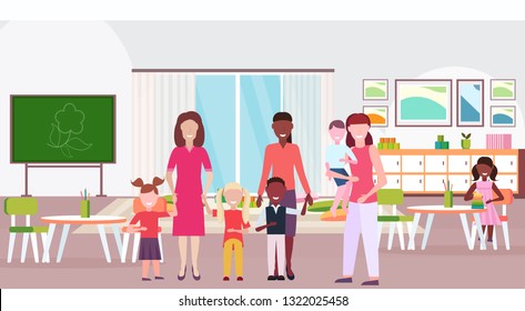 women teachers teaching mix race boys and girls preschool modern kindergarten children classroom with chalkboard desks chairs kid room interior full length flat horizontal