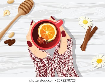 Women's hands wearing knitted gloves holding red cup of tea with different ingredients around