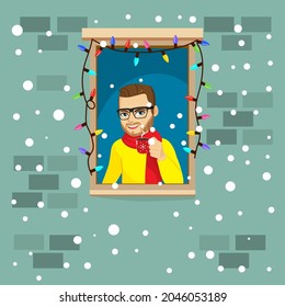 Young adult man with cup of hot chocolate looking through the window with colorful Christmas lights during winter snowfall