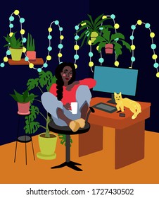 Young afro american woman sitting at desk with mug climbed on a chair with legs.Person works from home using computer and tablet,cat lies on table.Cozy room interior with plants in pots and lights.