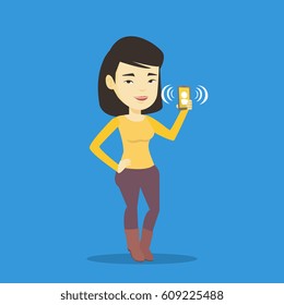 Young asian woman holding ringing mobile phone. Woman answering a phone call. Woman standing with ringing phone in hand. Vector flat design illustration isolated on blue background. Square layout.