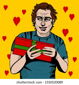 young attractive man in a blue T-shirt smiles and holds a gift in his hands in a red wrapper tied with a green ribbon on a yellow background with red hearts