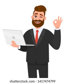 Young bearded business man is holding/showing a latest new laptop and gesturing/making okay or OK sign with hand fingers while winking eye. Modern technology, digital device/gadget concept in cartoon.