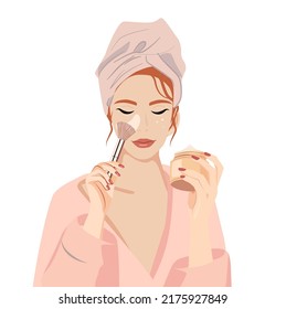 A young beautiful girl in a bathrobe and a towel on her head agnosticism cream on her face with a brush. The girl is holding a jar of cream in her hand. Conducts cosmetic procedures and cares for skin