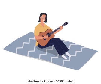 A young and beautiful girl sitting on a blanket and playing acoustic guitar