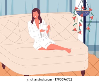 Young beautiful smiling woman sitting with bent legs on a large white comfortable sofa with cup of tea or drink, white bathrobe. Girl in the spa. Beauty treatments and relaxation. Cozy interior