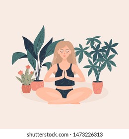 Young blonde woman in a black top sitting in a lotus position near the pots with tropical plants on a light background