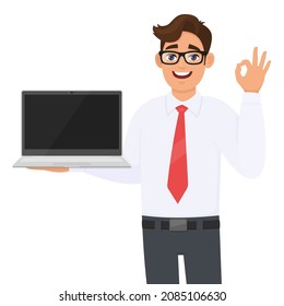 Young Business Man Showing Laptop And Gesturing Okay Or OK Sign. Handsome Person Carrying Computer. Male Character Holding Digital Technology Device. Cartoon  Illustration Design Concept In Vector.