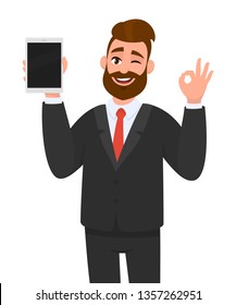 Young business man showing/holding blank screen of tablet computer display and gesturing/making okay, ok sign, while winking eye. Modern digital technology, gadget and device concept in cartoon style.