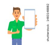 A young businessman is standing in a green shirt, smiling and showing a smartphone screen in his hand. The guy shows a thumbs up and holds a smartphone.