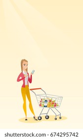Young caucasian woman standing near supermarket trolley full of products and holding a calculator in hand. Woman checking prices on the calculator. Vector flat design illustration. Vertical layout.