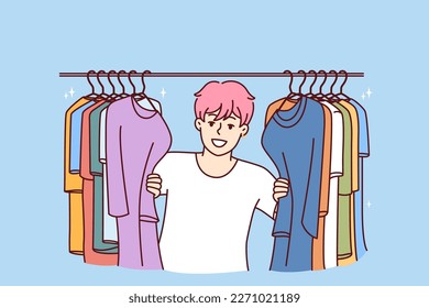 Young Cheerful man in t-shirt smiling peeking out from behind hanger for collection of clothes choosing new look. Guy with pink hair is standing in clothing store wanting to buy fashionable outfit 