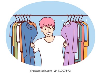 Young Cheerful man in t-shirt smiling peeking out from behind hanger for collection of clothes choosing new look. Guy with pink hair is standing in clothing store wanting to buy fashionable outfit