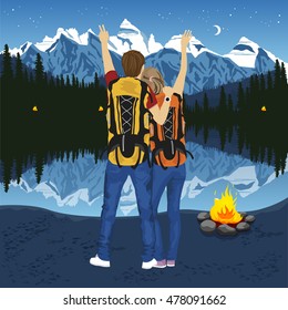 young couple of hikers with hands up enjoying mountain lake at night near campfire