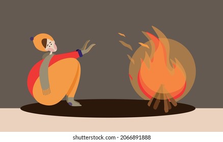 A young guy, boy in a yellow hat and a green scarf, a red sweater and orange pants sits outdoor and warms his hands on a fire or campfire in a children's camp in cold autumn or winter weather. 