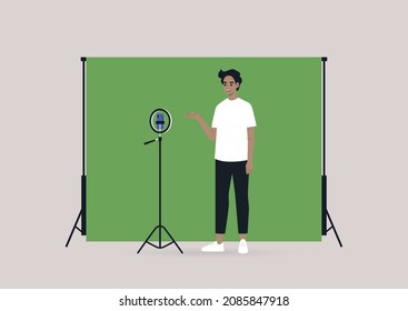 A young male Caucasian blogger recording a video with a tripod, a mobile phone, and a chroma key screen