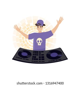 Young male DJ in purple cap and t-short stands with raised arms at mixing consol on yellow light chaser