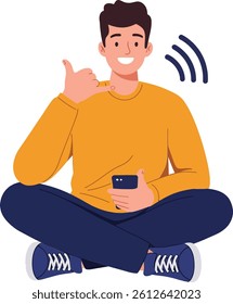 A young man with a friendly smile sits cross-legged, holding a smartphone while gesturing with a thumbs-up, conveying a positive and relaxed mood.