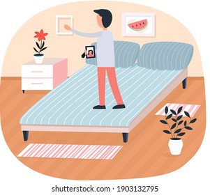 Young man householder puts things in order at home standing on bed in bedroom insert photo into frame on wall. Stay at tidy home. Family members together activity for order things in their places