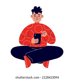 A young man in a red sweater sits on the floor and works with the phone on social networks.