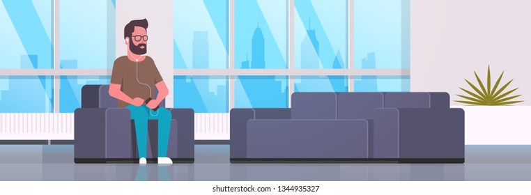 young man sitting armchair listening to music on smartphone with headphones  