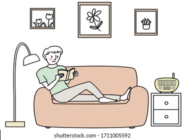 Young man sitting on couch and reading book comfortably. Cute happy man reading book on sofa. Cheerful man enjoying leisure time reading book.