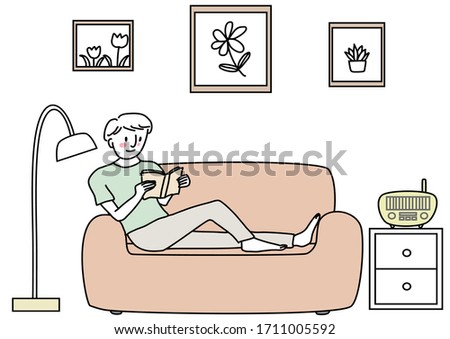Young man sitting on couch and reading book comfortably. Cute happy man reading book on sofa. Cheerful man enjoying leisure time reading book.
