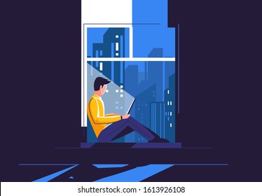 A young man is sitting on the windowsill with a laptop, in the dark against the background of the night city outside the window