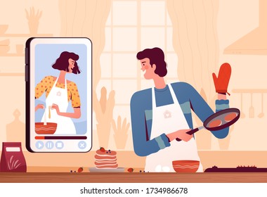 A young man watches a video recipe on the phone and prepares pancakes at home in the kitchen. The concept of online recipes and home cooking