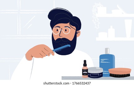 Young man wearing glasses brushing his beard and looking in the mirror having styling cosmetic, brushes and odecolone bottle in his bathroom. Everyday morning routine. Men hygiene and self care.