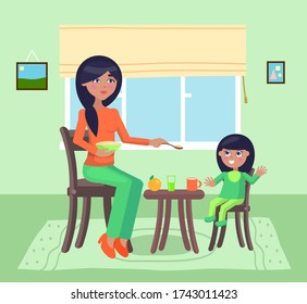 Young mother feeding her daughter give spoon with poridge or meal sitting at table. Little girl smiling and have fun, rejoice. Lunch, dinner or supper time. Apple, glass, cup of drink at table