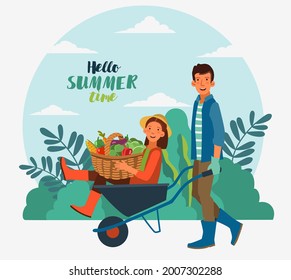 Young and smiling man pushing his laughing girlfriend in a wheelbarrow. Cute woman in a garden with a basket full of fresh vegetables: cabbage, pepper, tomato, carrot, corn, broccoli and eggplant 